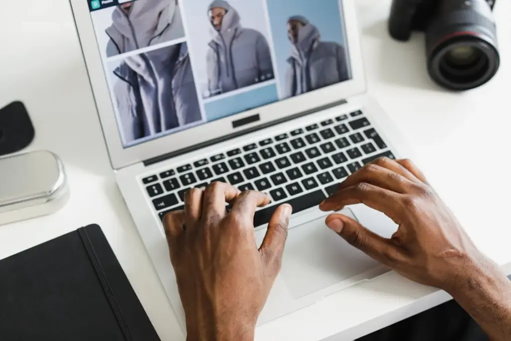 Person typing on a laptop showing images of a jacket, with a camera and a notebook nearby on a white desk.