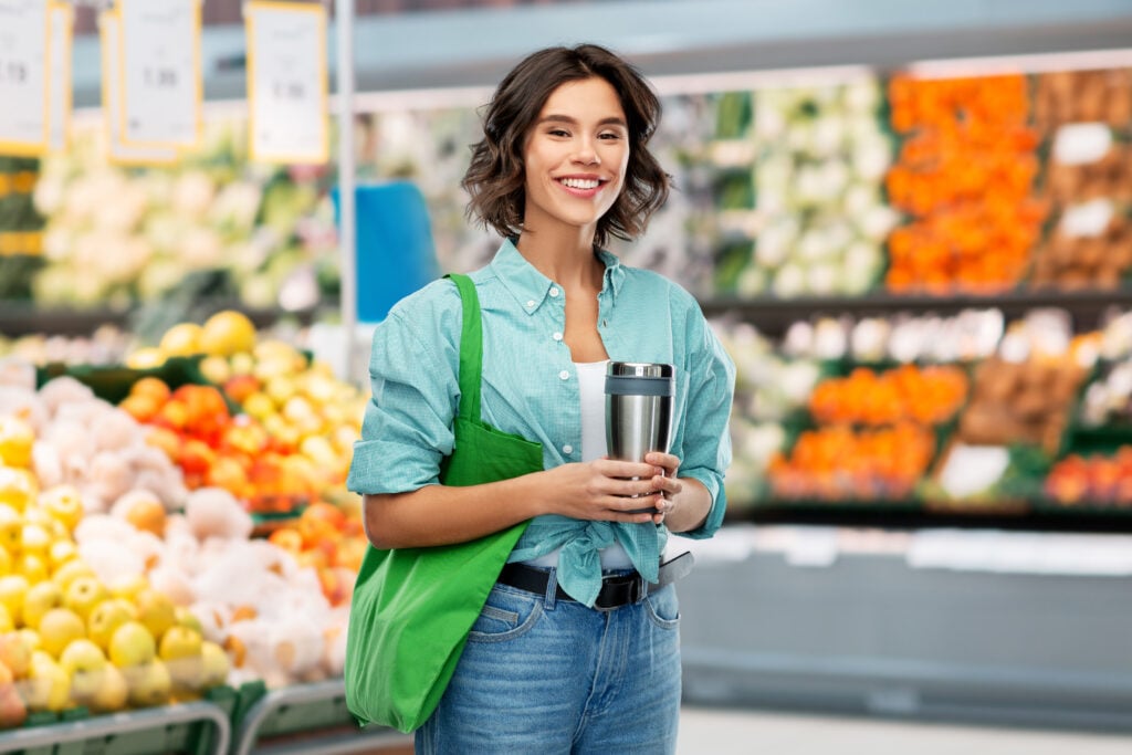 A woman holding a reusable cup and carrying a green tote bag stands in a grocery store, with shelves of fresh produce in the background.