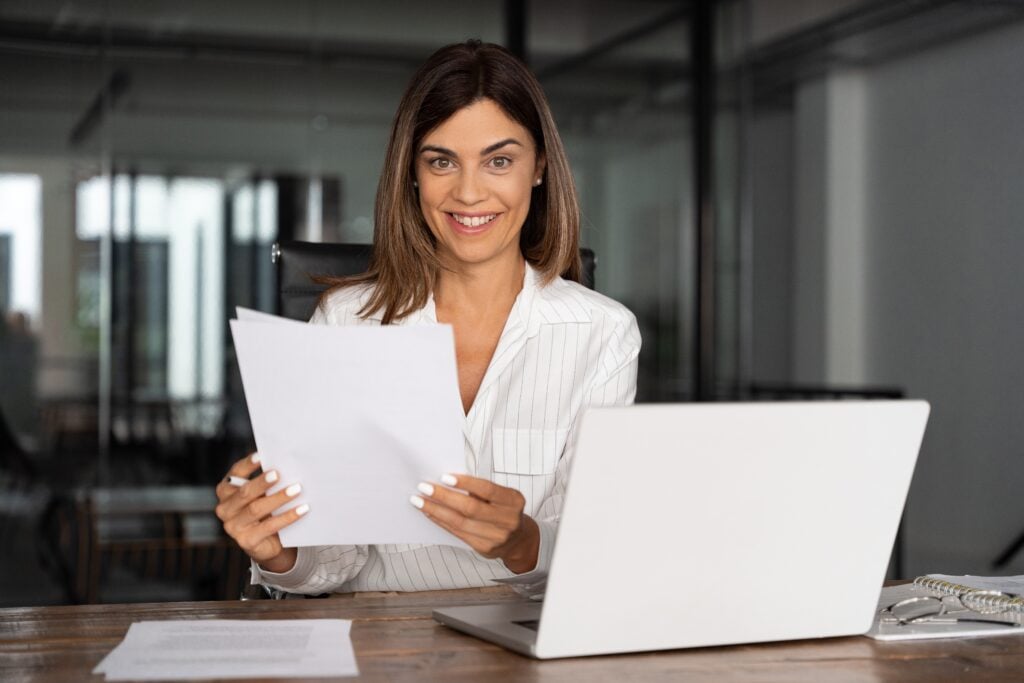 A woman in a white shirt sits at a desk holding papers, smiling, with a laptop open in front of her.