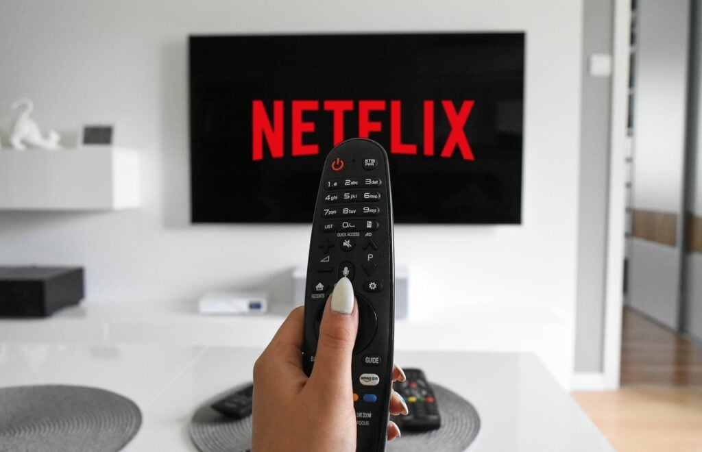 A hand holding a remote control points at a TV displaying the Netflix logo.