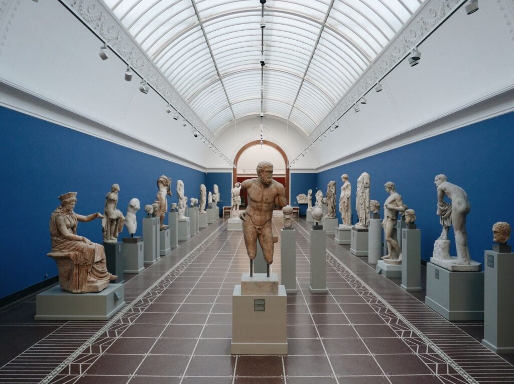 A museum hall displaying numerous ancient sculptures on pedestals, with a high arched ceiling and blue walls.