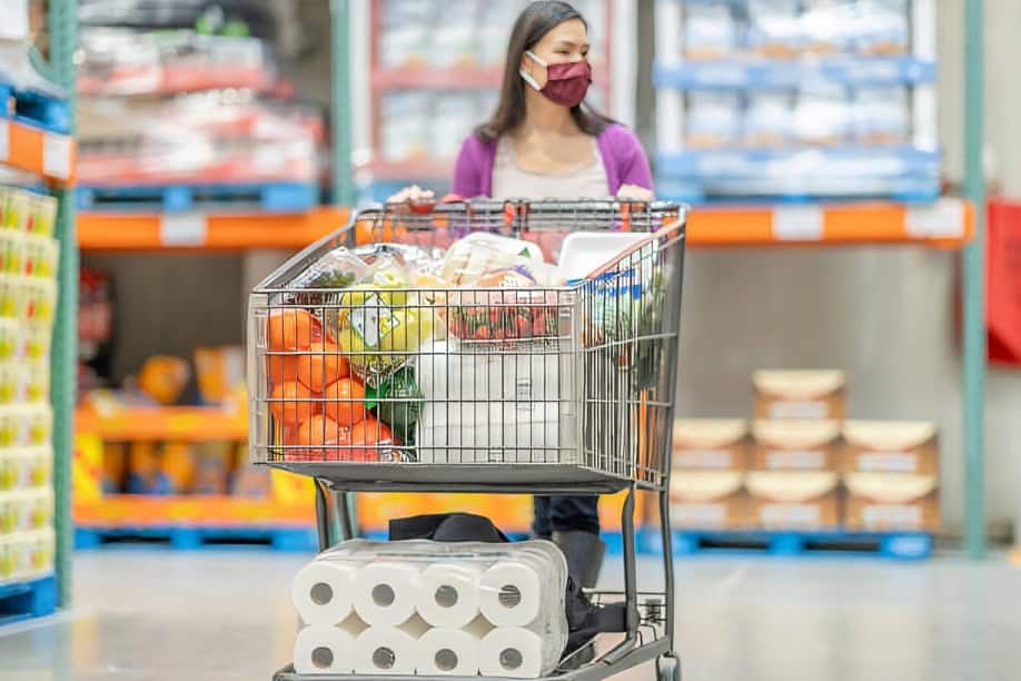 A woman wearing a mask pushes a shopping cart filled with groceries and toilet paper in a warehouse store.