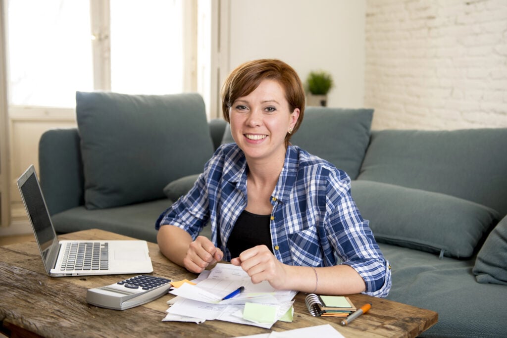 Person sitting at a table with a laptop, papers, and a calculator, smiling at the camera. Sofa and window in the background.