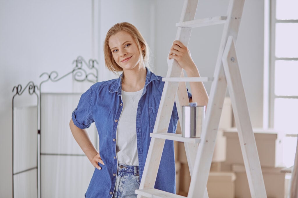 A person in a blue shirt stands next to a stepladder, holding a can of paint in a bright room.