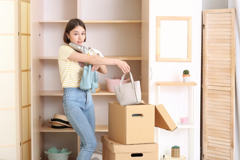 A woman organizing a room, holding several handbags and standing next to cardboard boxes in front of empty shelves.