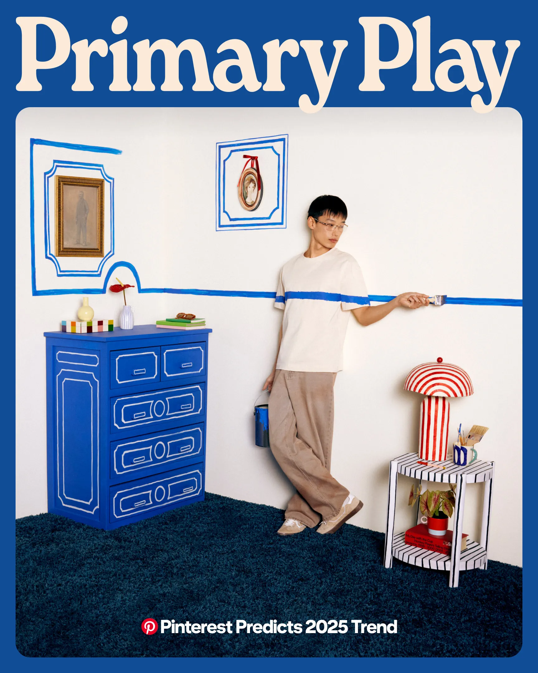 A man stands against a white wall in a white shirt and baggy beige pants. He holds a paintbrush, and there’s a blue stripe across his shirt and the wall. The room contains a blue dresser, a red-striped lamp and a blue-striped side table.