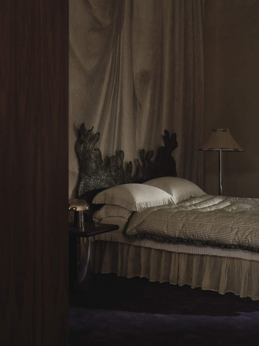 A ruffle bed skirt on bed in a moody and dark bedroom. There is a custom, abstract stone headboard, a deep purple carpet, and drapery hanging on the wall behind the bed