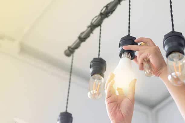 Hands screwing a light bulb into a ceiling fixture with several hanging light bulbs.