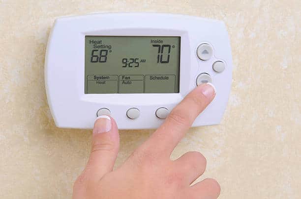 A hand adjusts the settings on a white digital thermostat mounted on a beige wall. The display shows a heat setting of 68°F, an inside temperature of 70°F, and the time as 9:25 AM.