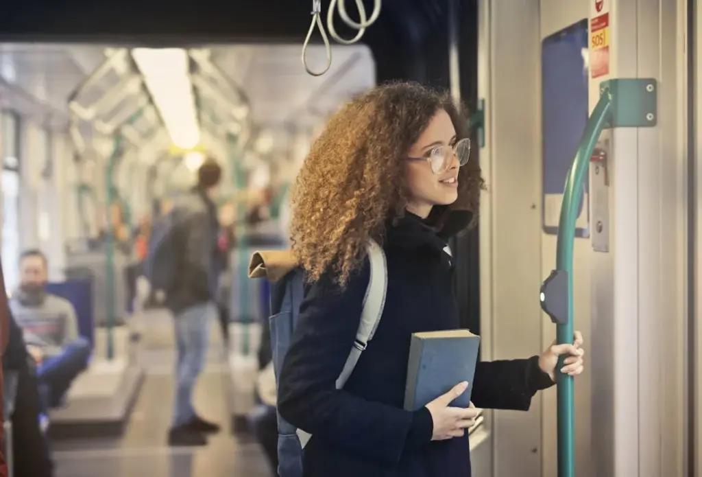 A person with curly hair and glasses holds a book and stands by the door inside a train, while other passengers are visible in the background.